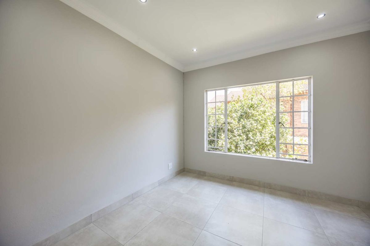 A bright, empty bedroom featuring neutral tiled flooring, recessed downlighting, and a large window with security bars.
