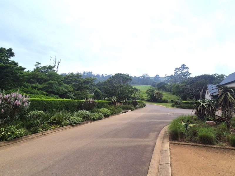 A well-maintained paved road leading through a landscaped residential estate with mature trees and lush greenery.
