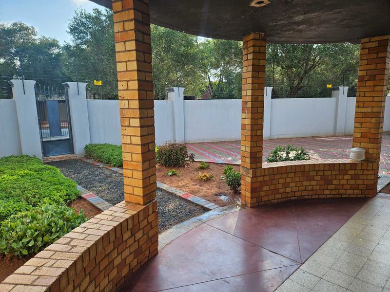 A covered patio area featuring brick pillars and flooring, overlooking a paved driveway and secure perimeter wall with electric fencing.