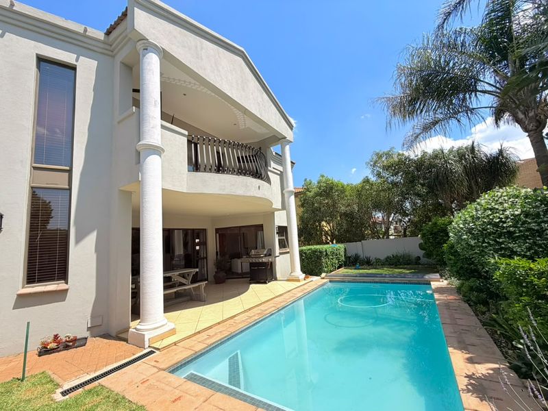 A well-maintained outdoor area featuring a swimming pool, a covered patio with classical columns, and an upper-level balcony overlooking the garden.