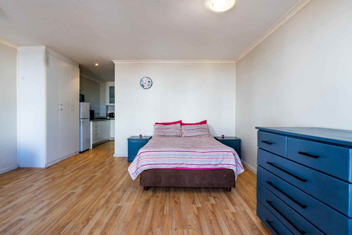 A spacious bedroom featuring light wood-look flooring, built-in cupboards, and an open-plan layout leading to a compact kitchen area.