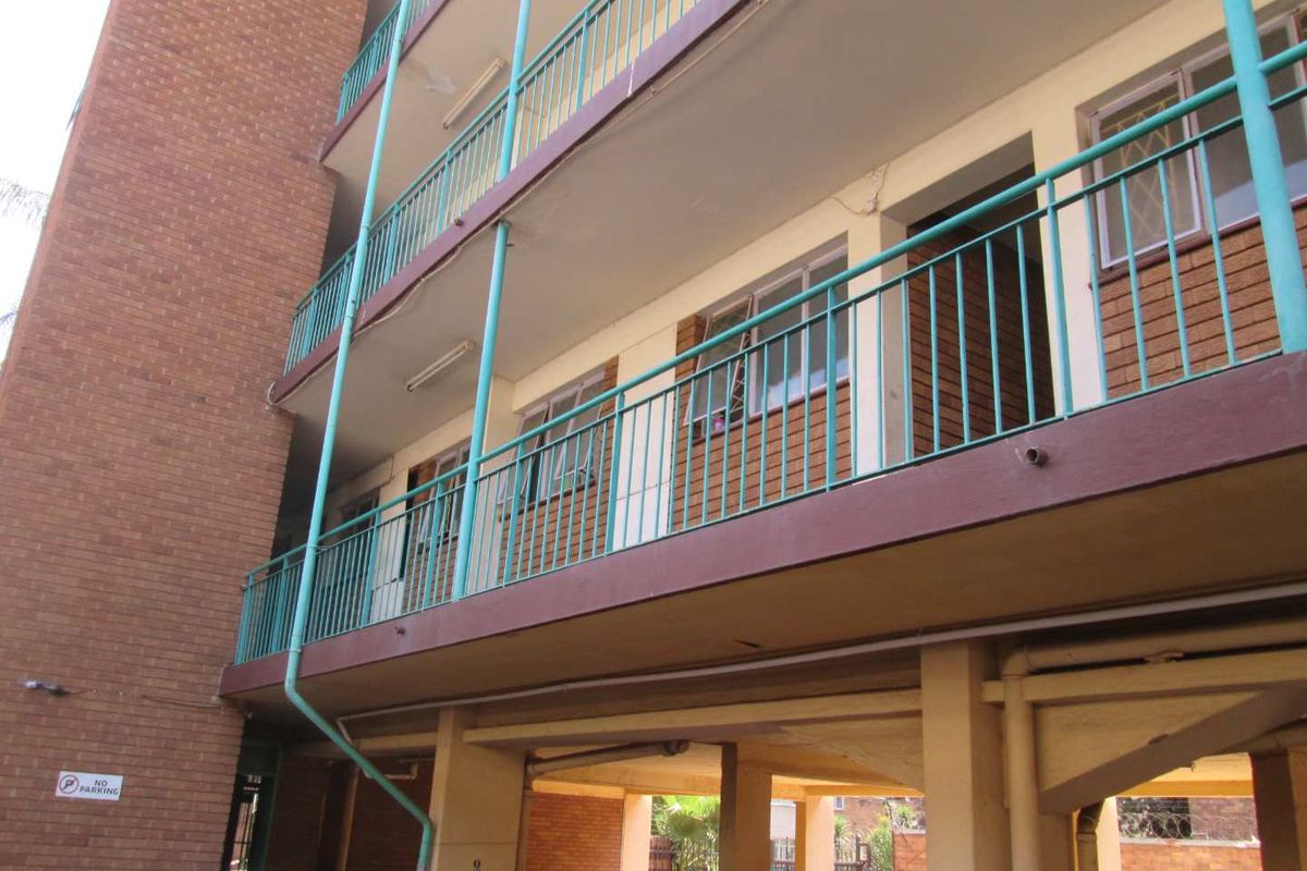 Exterior view of an apartment building featuring brick facade, covered walkways with metal railings, and ground-level parking area.