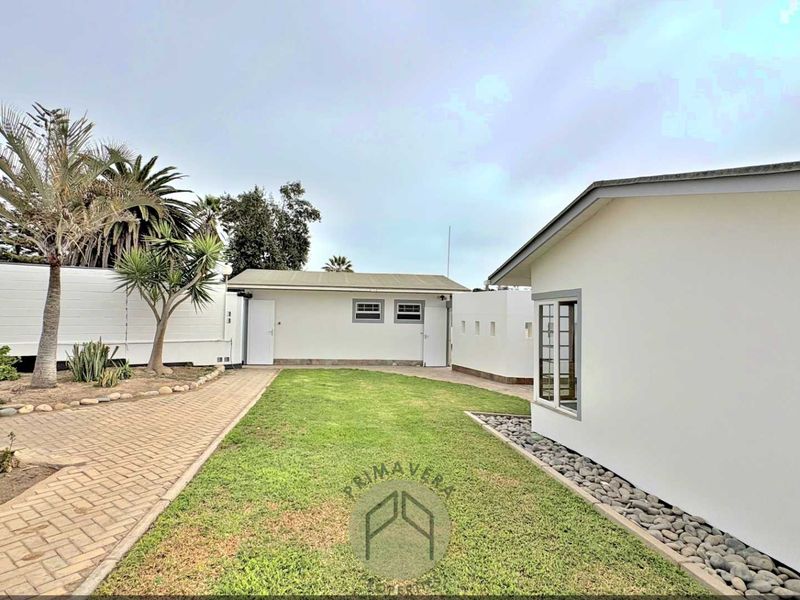 A well-maintained rear garden area featuring a paved walkway, a central lawn, and a detached outbuilding with white exterior walls.