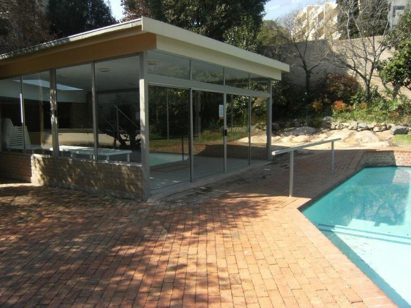 Outdoor pool area featuring brick paving, a metal handrail, and an adjacent glass-enclosed structure set against a landscaped retaining wall.