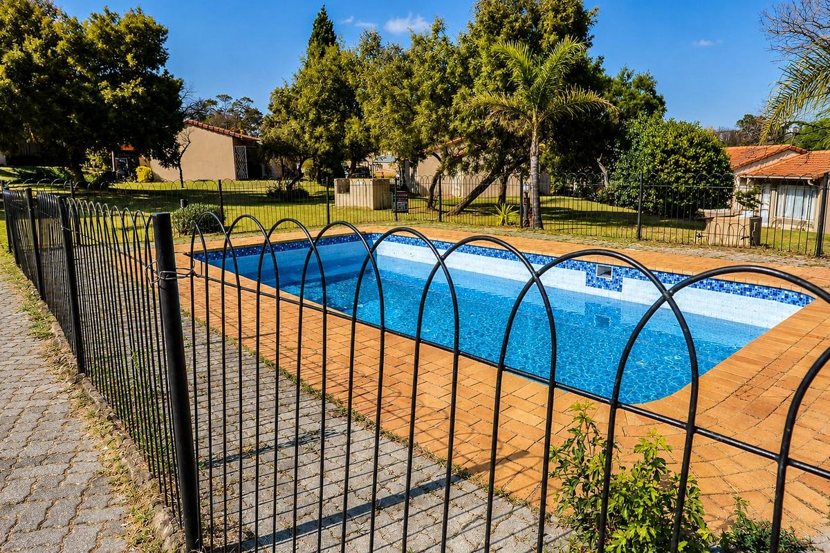 A well-maintained outdoor swimming pool area featuring a paved surround, secure metal perimeter fencing, and established garden landscaping within the townhouse complex.