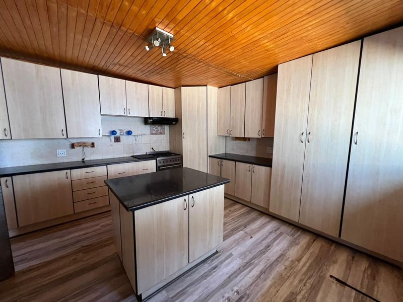 A functional kitchen featuring light wood-tone cabinetry, a central island, dark countertops, and a timber-clad ceiling, offering a clean and practical workspace.
