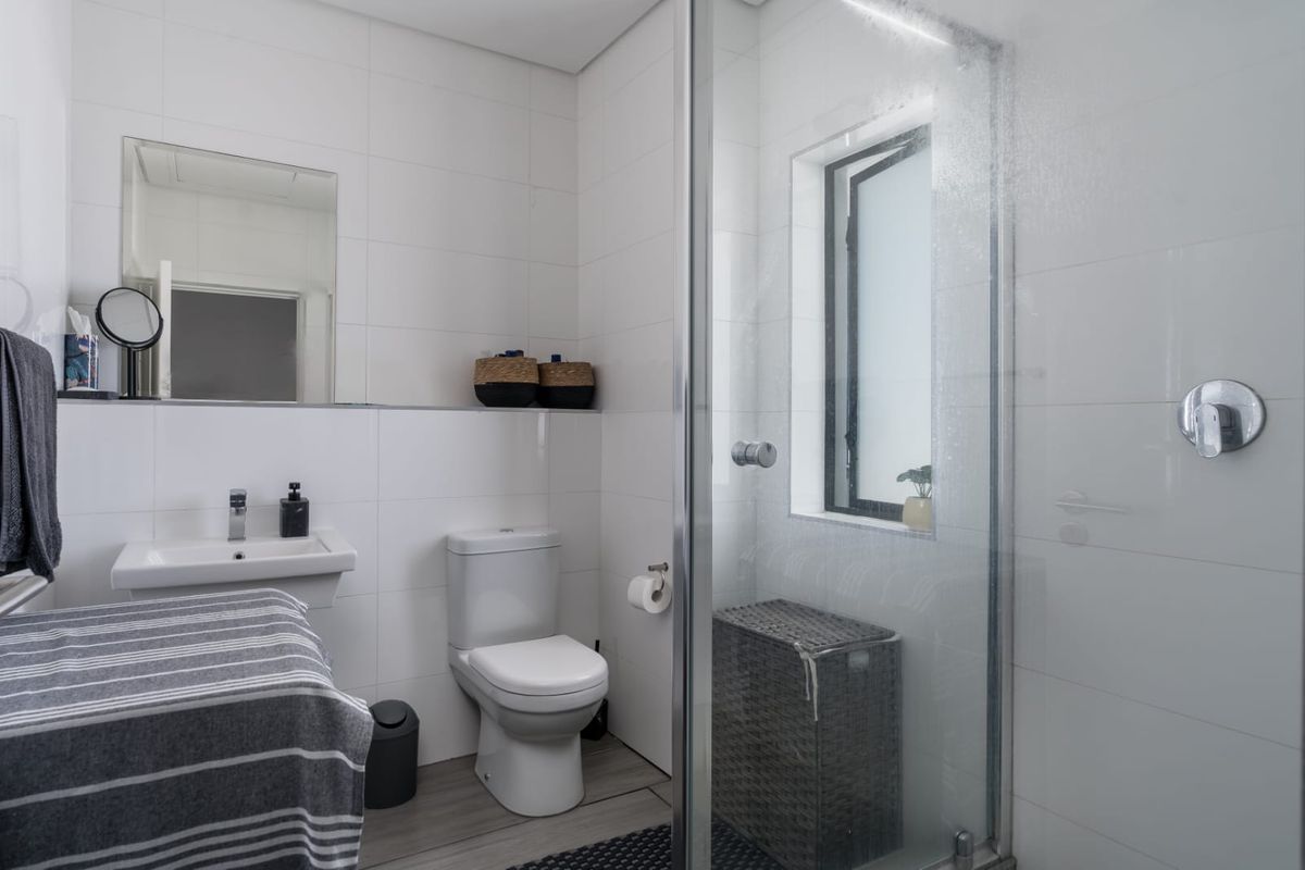 A clean, modern bathroom featuring a wall-mounted basin, toilet, and a glass-enclosed shower with white wall tiles and neutral floor tiling.