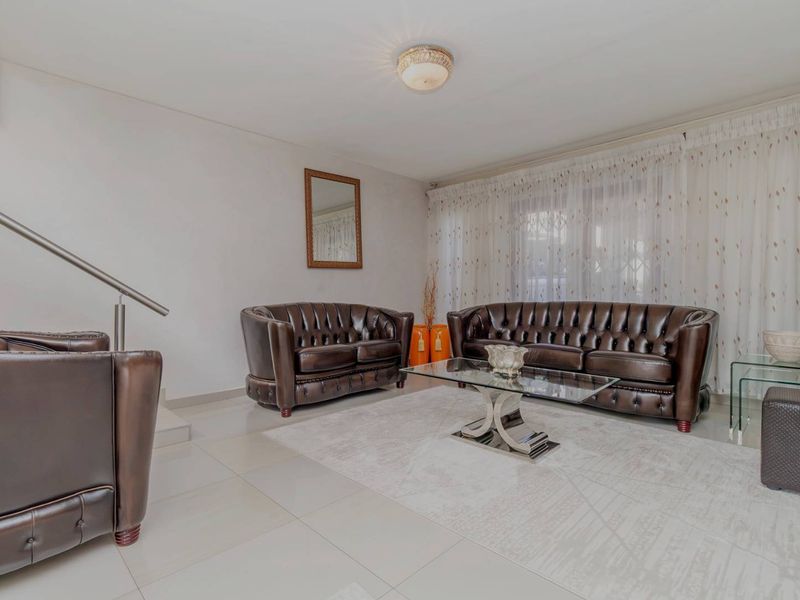 A well-maintained living room featuring neutral tiled flooring, a modern stainless steel handrail, and ample natural light through curtained windows.