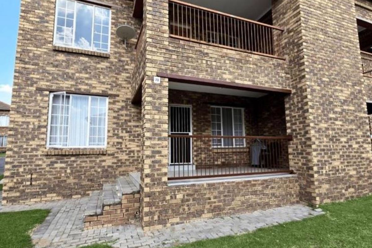 A multi-story apartment building exterior featuring face brick construction, a ground-floor patio, and an upper-level balcony with metal railings.