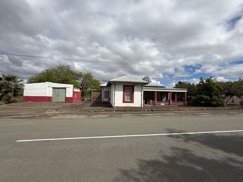 Front view of a single-story house featuring a covered veranda, corrugated metal roof, and a separate detached garage structure on a paved lot.