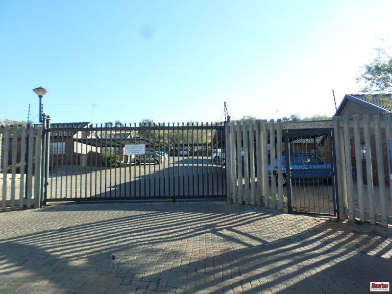 Secure gated entrance to the apartment complex featuring a paved driveway, perimeter wall with electric fencing, and covered parking areas.