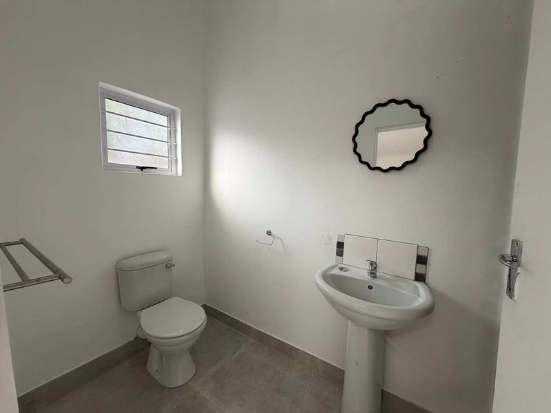 A functional, empty bathroom featuring a pedestal washbasin, toilet, tiled flooring, and a window with security bars.