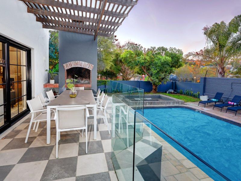 Covered outdoor patio featuring a built-in braai, glass balustrade, and direct access to a swimming pool and landscaped garden area.