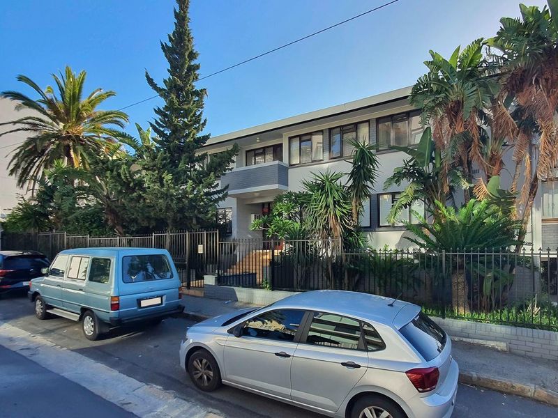 Street-level view of the apartment building facade featuring a concrete exterior, metal perimeter fencing, and established garden landscaping.