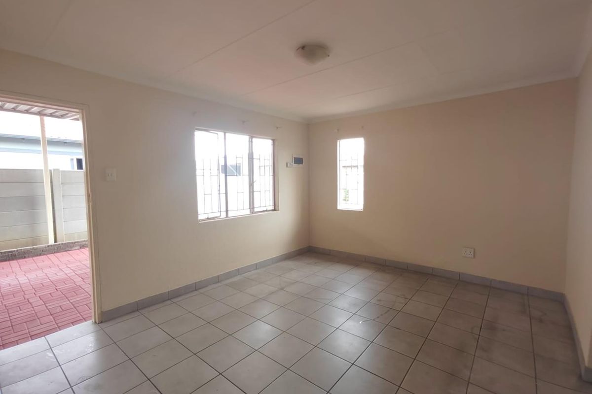An empty living room featuring neutral tiled flooring, security bars on the windows, and direct access to an outdoor paved area.