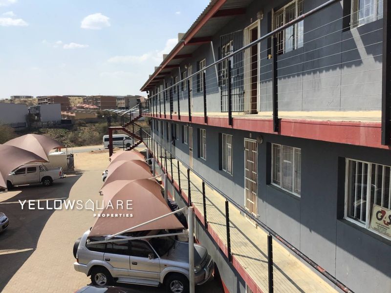 Exterior view of a multi-story apartment complex featuring a metal walkway, security gates, and covered parking bays.