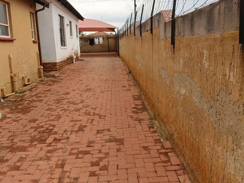 A side exterior view of the property featuring a paved walkway, a high perimeter wall with security razor wire, and a shade sail structure.