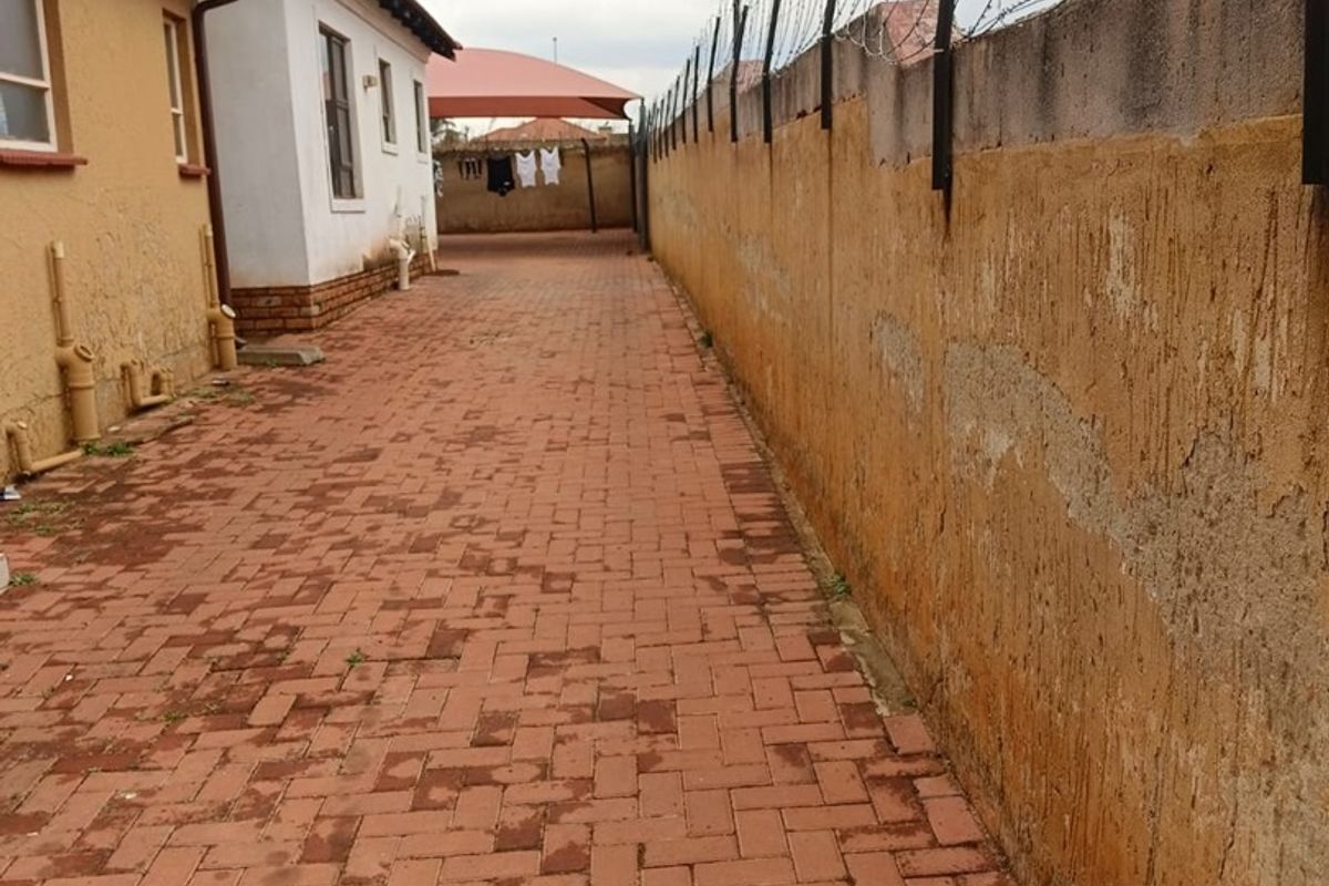 A side exterior view of the property featuring a paved walkway, a high perimeter wall with security razor wire, and a shade sail structure.