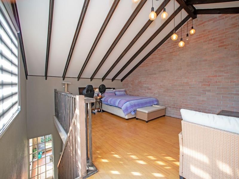 A spacious loft-style bedroom featuring exposed timber beams, a striking brick feature wall, and warm laminate flooring, overlooking the lower level.