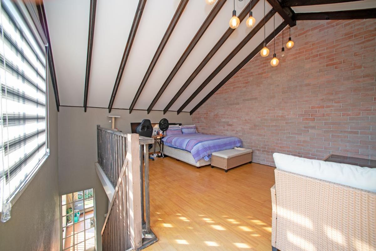 A spacious loft-style bedroom featuring exposed timber beams, a striking brick feature wall, and warm laminate flooring, overlooking the lower level.