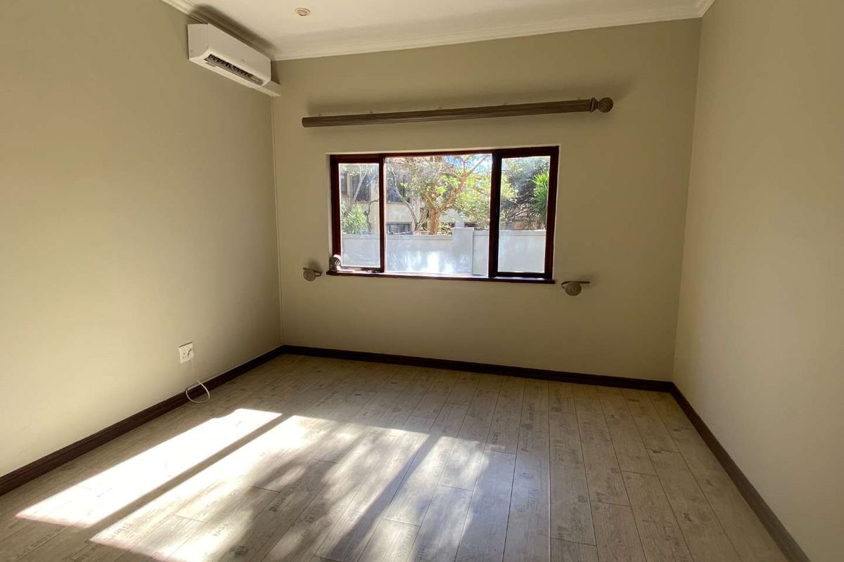 A bright, empty bedroom featuring wood-look laminate flooring, a wall-mounted air conditioning unit, and a window with curtain rod hardware.