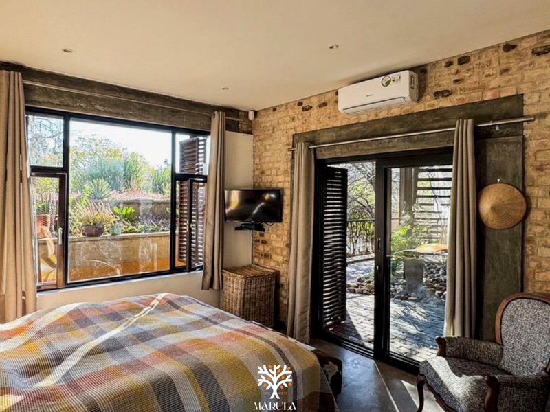 A well-lit bedroom featuring exposed brick walls, concrete flooring, and large sliding glass doors that provide direct access to an outdoor patio area.