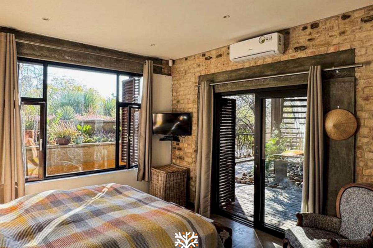 A well-lit bedroom featuring exposed brick walls, concrete flooring, and large sliding glass doors that provide direct access to an outdoor patio area.