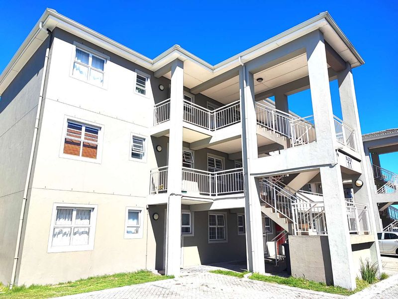 A multi-story apartment building exterior featuring balconies with metal railings, an external staircase, and a paved driveway area.