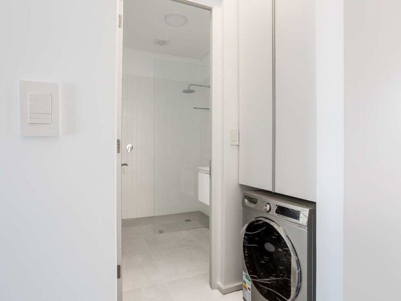 A clean, modern laundry area featuring built-in cabinetry and tiled flooring, with a direct view into a contemporary bathroom with a glass shower enclosure.