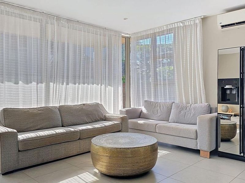 A bright and spacious living room featuring large windows with sheer curtains, tiled flooring, and a wall-mounted split-system air conditioner.