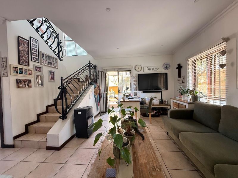 Spacious living room featuring tiled flooring, a decorative wrought iron staircase railing, and natural light through large windows with venetian blinds.