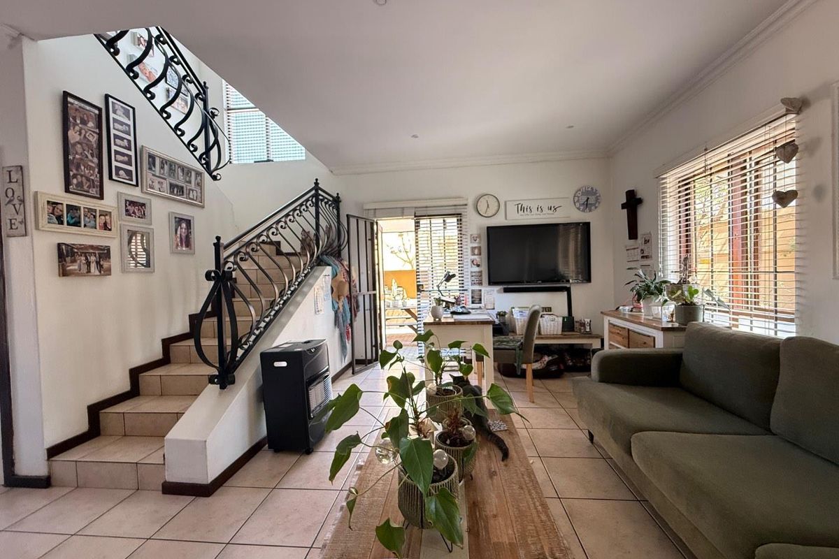 Spacious living room featuring tiled flooring, a decorative wrought iron staircase railing, and natural light through large windows with venetian blinds.