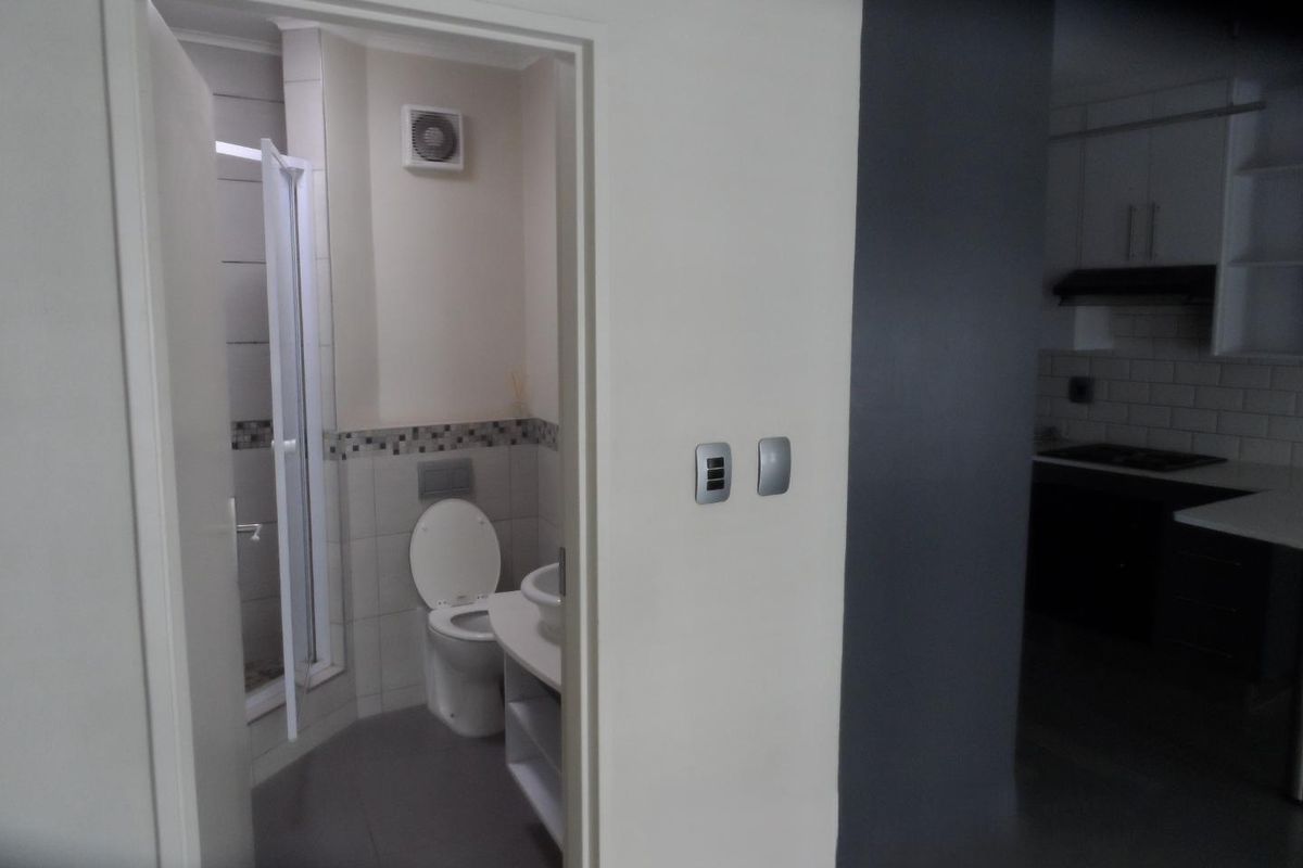A compact bathroom featuring a shower, toilet, and vanity, adjacent to a kitchen area with dark cabinetry and subway tile splashback.