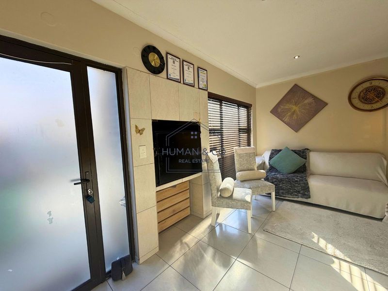 A bright living room featuring tiled flooring, a built-in media unit, and a frosted glass entrance door with natural light filtering through venetian blinds.