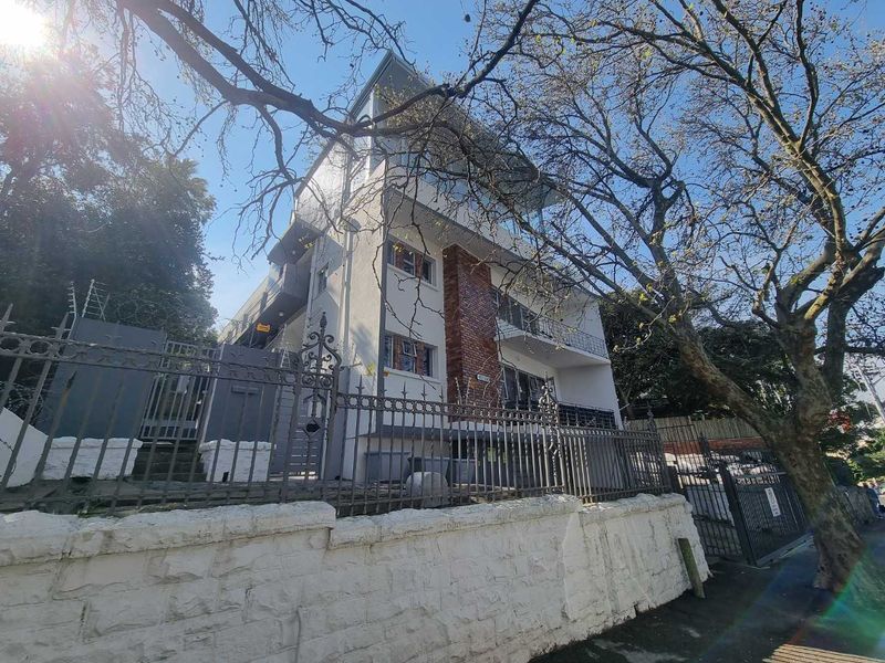 Exterior view of a multi-story apartment building featuring balconies, brick accents, and a secure wrought iron perimeter fence with a stone retaining wall.