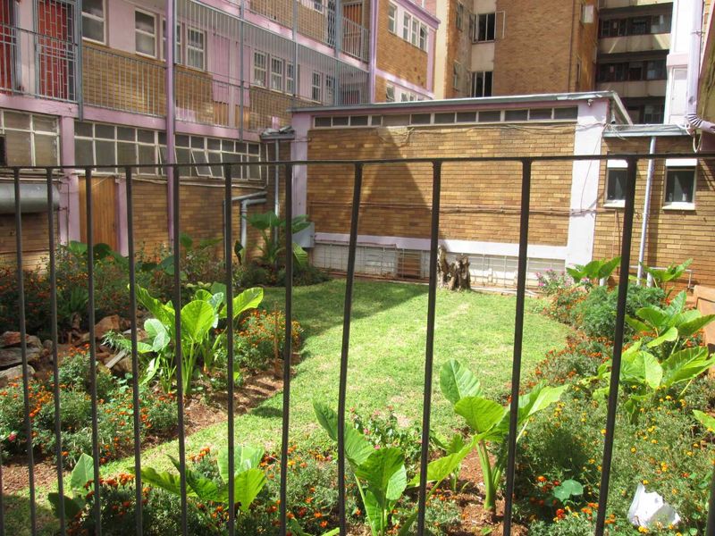 A rear courtyard garden area featuring a lawn, established garden beds, and metal security fencing, set against a brick apartment building facade.