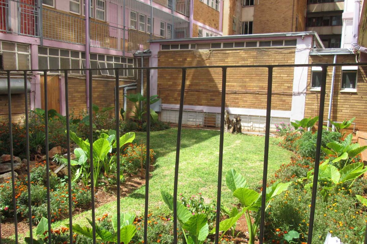 A rear courtyard garden area featuring a lawn, established garden beds, and metal security fencing, set against a brick apartment building facade.