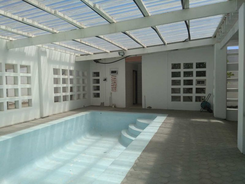An enclosed indoor swimming pool area featuring a translucent roof, concrete block ventilation screens, and paved flooring, currently in need of maintenance.