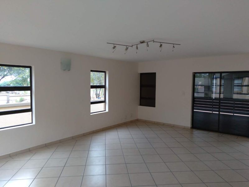 Spacious, empty living area featuring neutral tiled flooring, modern track lighting, and large windows providing ample natural light with access to an outdoor area.
