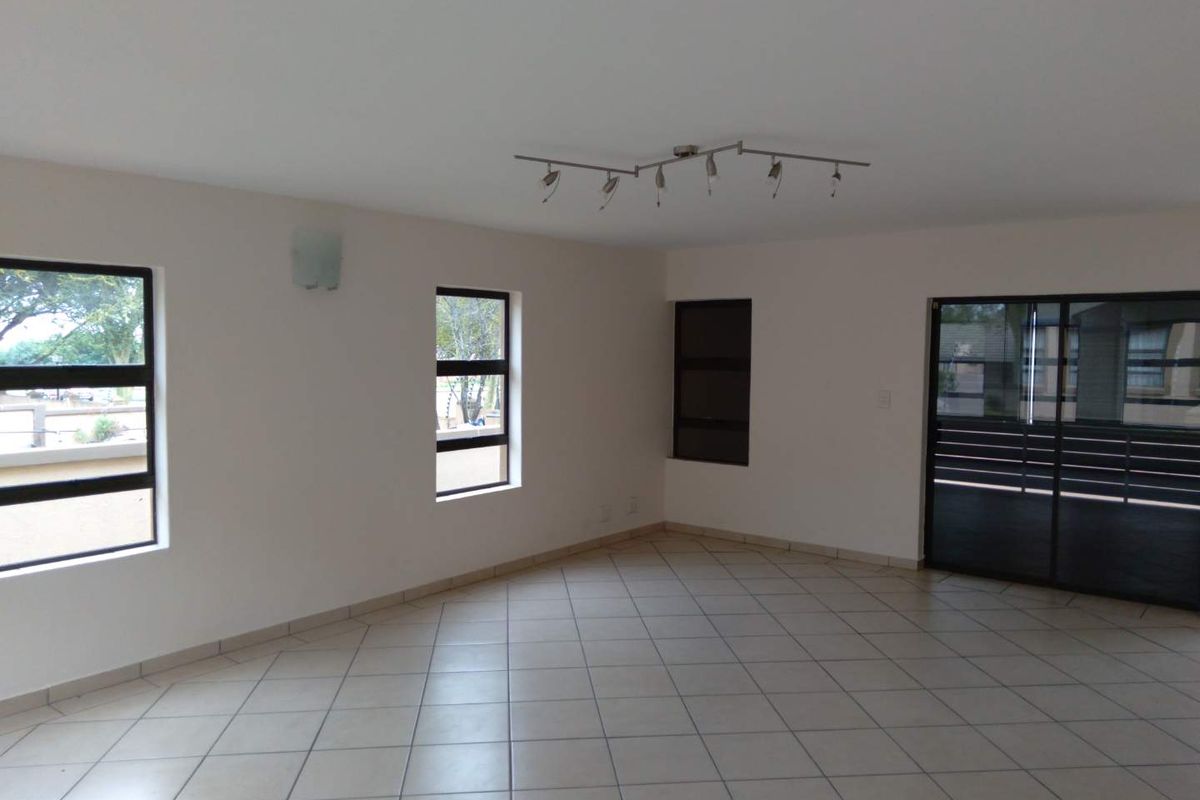 Spacious, empty living area featuring neutral tiled flooring, modern track lighting, and large windows providing ample natural light with access to an outdoor area.