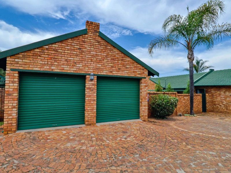 A double garage with green roller doors and face brick facade, featuring a circular paved driveway and established garden surroundings.