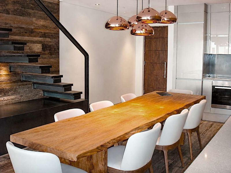 Modern open-plan dining area featuring a striking floating staircase, timber wall accents, and a sleek integrated kitchen with high-end finishes.