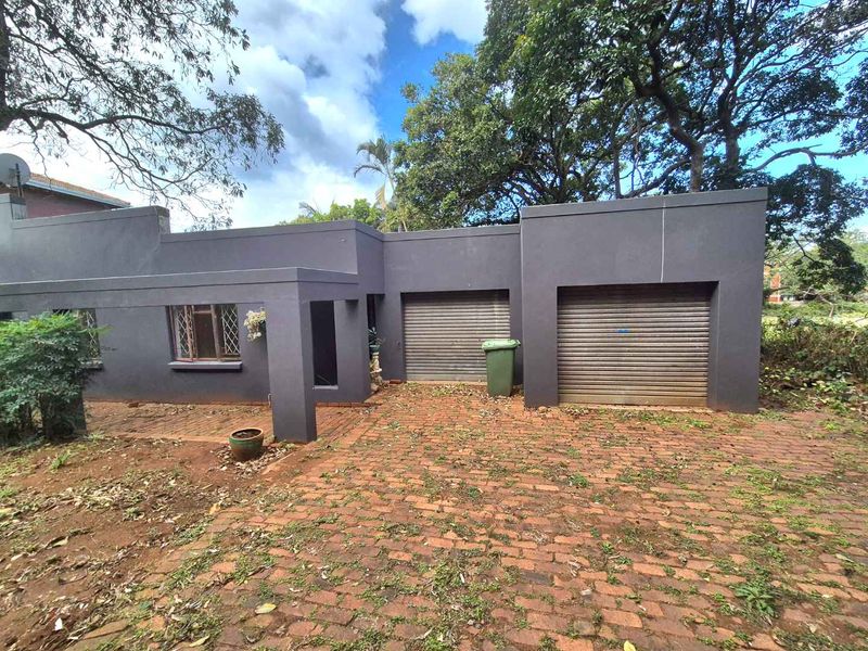 Front exterior view of the property featuring a double garage with roller shutter doors and a brick paved driveway surrounded by mature trees.