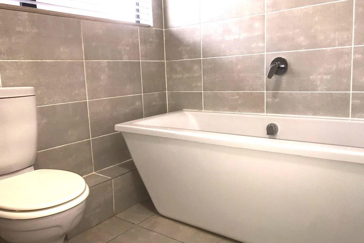 A modern bathroom featuring neutral wall tiles, a built-in bathtub with wall-mounted chrome fixtures, and a standard toilet.