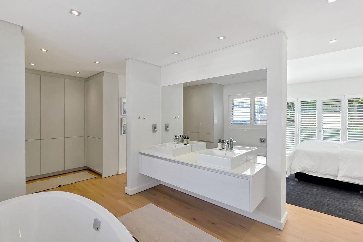 Modern open-plan bathroom featuring a double vanity, freestanding bathtub, hardwood flooring, and built-in cabinetry with integrated lighting and plantation shutters.