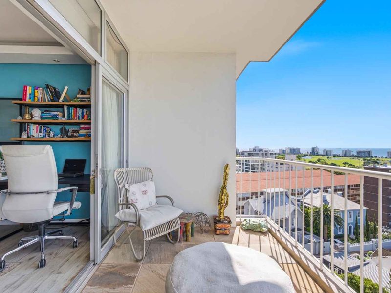 A bright balcony featuring tiled flooring and a metal balustrade, offering expansive urban and coastal views, seamlessly connected to an interior office space.