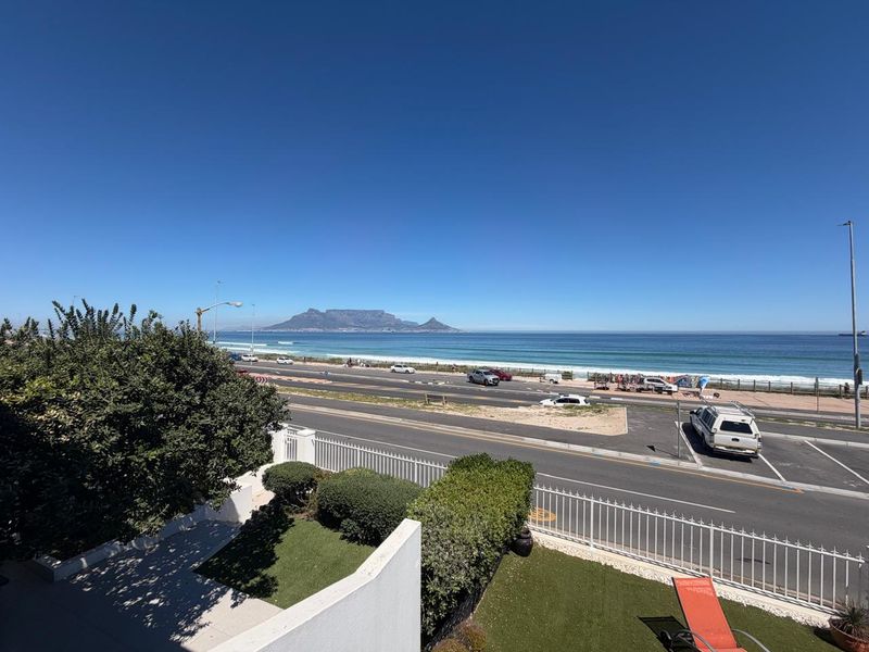A scenic elevated view overlooking the coastal road, beach, and Table Mountain in the distance, featuring well-maintained perimeter fencing and garden space.