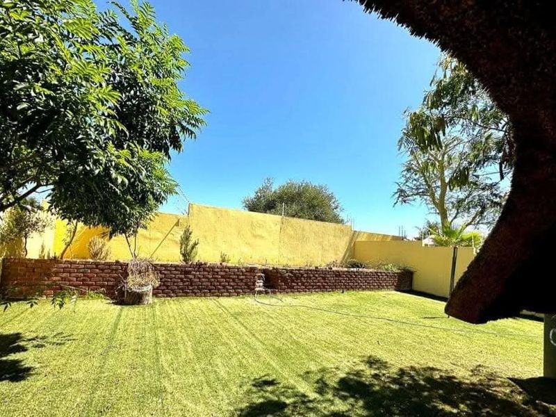 A private backyard garden featuring a manicured lawn, a brick retaining wall, and a perimeter boundary wall, viewed from under a thatched roof patio.