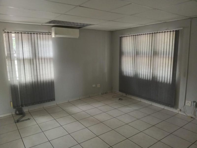 An empty office space featuring ceramic floor tiles, a suspended ceiling, and a wall-mounted air conditioning unit with vertical window blinds.