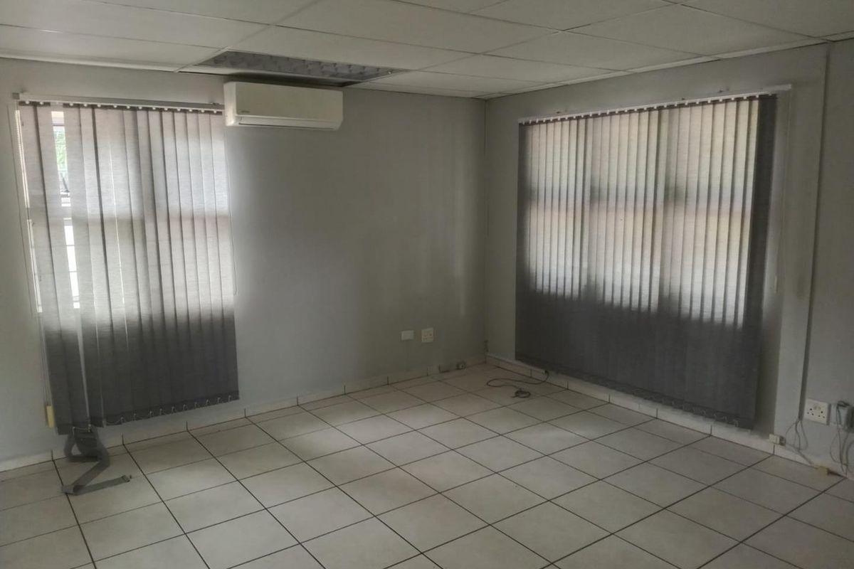An empty office space featuring ceramic floor tiles, a suspended ceiling, and a wall-mounted air conditioning unit with vertical window blinds.